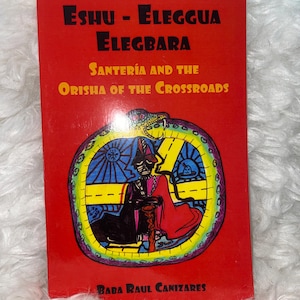 May include: A red book titled "Eshu - Eleggua Elegbara" with the subtitle "Santería and the Orisha of the Crossroads." The cover features an illustration of a figure within a colorful, circular design. The author's name, Baba Raul Canizares, is at the bottom.