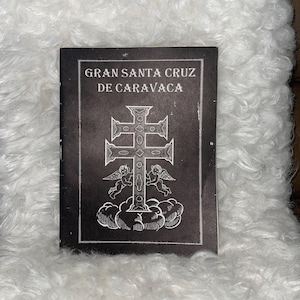 May include: A black booklet with the title "GRAN SANTA CRUZ DE CARAVACA" in white lettering. The cover features a detailed illustration of a cross with angels and clouds. The booklet is resting on a white, fluffy surface.