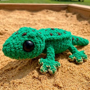 Può includere: Un geco verde all'uncinetto fatto a mano con macchie marroni e grandi occhi neri. Il geco è adagiato su un letto di sabbia. I piedi del geco sono di una tonalità di verde più chiara.