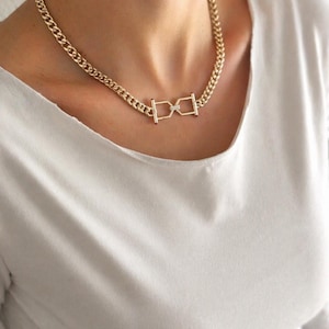Pode incluir: Colar de corrente dourada com um pingente geométrico único. O colar apresenta uma corrente grossa e um pingente central com um detalhe em forma de diamante. A joia é usada sobre uma blusa branca.
