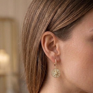 Puede incluir: Pendientes de aro dorados con diseño de brújula. Los pendientes presentan un colgante circular con un diseño de estrella y están sujetos a un aro pequeño. Los pendientes son de un material metálico brillante.