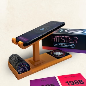 Pode incluir: Um suporte de plástico laranja segurando um smartphone, acompanhado por uma pilha de fichas de jogo circulares pretas com o texto HITSTER, uma caixa de jogo rotulada HITSTER DAS MUSIK-PARTY SPIEL e dois cartões de jogo quadrados com 1985 e 1988.