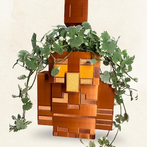 Può includere: Un vaso di colore rame, squadrato e pixelato, a forma di personaggio in stile Minecraft con testa e corpo quadrati. Il vaso presenta occhi e naso arancioni brillanti, con una pianta ricadente variegata di colore verde e bianco che cresce.