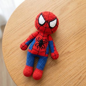 May include: A handmade crochet doll featuring the iconic red and blue superhero suit. The head is round with large white eyes outlined in black, and the torso displays a black spider emblem. The limbs are red and blue with detailed stitching.