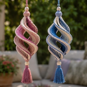 May include: Two hanging spiral ornaments crafted from crocheted yarn. One ornament features a gradient of pink and cream, while the other displays blue and cream. Each piece is topped with a wooden bead and finished with a matching yarn tassel.