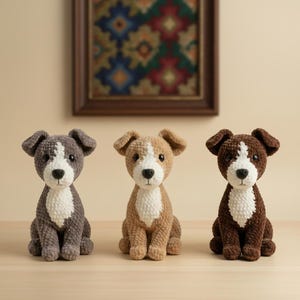 May include: Three crocheted dog figurines in a row. The dogs are in shades of gray, tan, and brown, each with a white chest and face markings. They have floppy ears and are sitting upright. A framed artwork hangs in the background.
