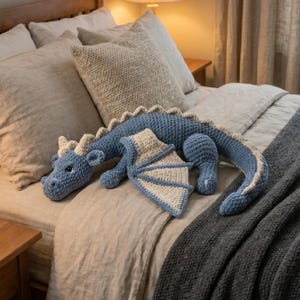 Può includere: Un peluche a forma di drago blu e crema all'uncinetto con un corpo lungo, ali e corna. Il drago è adagiato su un letto con biancheria da letto dai colori neutri e una coperta grigia. Il corpo del drago è lungo circa 76 cm.