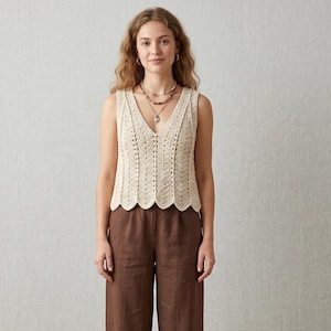 Puede incluir: Un top de ganchillo sin mangas de color crema con un escote en V profundo y un dobladillo festoneado. La prenda está confeccionada con paneles verticales de intrincado calado y se combina con pantalones de color marrón oscuro.