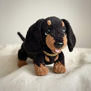 Puede incluir: Un perro salchicha de ganchillo hecho a mano en negro y marrón. El perro tiene un collar de cadena dorada y una lengua rosa. El perro posa sobre una superficie de color neutro.