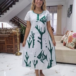 May include: White dress with a green fish and coral pattern. The dress has short sleeves and a round neckline with green trim. The design features stylized fish and coral motifs in a dark green color, creating a nautical theme. The dress falls to the mid-calf.