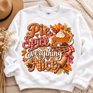 Puede incluir: Una sudadera blanca de cuello redondo con un estampado gráfico colorido con el texto Pie, Spice & Everything Nice. El diseño incluye una porción de tarta de calabaza con nata montada, un rodillo de madera, canela y hojas de otoño.