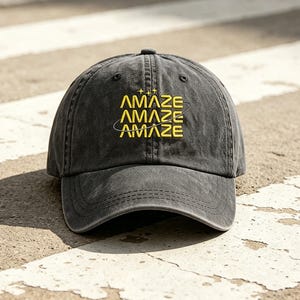 May include: A charcoal gray baseball cap with the word "AMAZE" in yellow, stacked three times. The cap has a curved brim and a classic six-panel design. The text has a star and planet design.