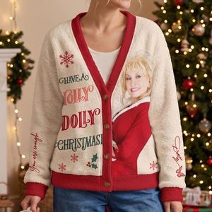 Puede incluir: Un cárdigan de forro polar color crema con ribetes rojos, que presenta un gráfico de Dolly Parton con un conjunto rojo. El texto dice Have a Holly Dolly Christmas, con detalles de copos de nieve y acebo. Las mangas incluyen el texto You are enough y Dolly.