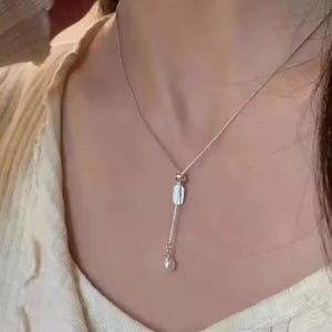 May include: A silver-toned lariat necklace featuring a delicate box chain, a small rectangular metal tag with a textured surface, and a single round white pearl pendant suspended at the end of the adjustable drop chain.