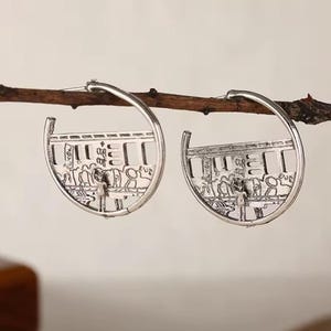 May include: A pair of silver-toned hoop earrings. Each earring features a detailed, openwork design depicting a scene with figures and architectural elements. The earrings are suspended from a brown twig, showcasing their intricate craftsmanship and unique style.