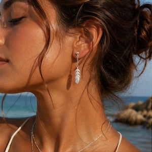 May include: Silver feather earrings. Each earring features a small, detailed feather pendant suspended from a delicate chain. The earrings are shown in a close-up shot, highlighting their intricate design and the way they catch the light.