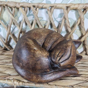 May include: A dark brown wooden carving of a curled-up sleeping cat. The sculpture features smooth, rounded contours and carved lines that define the cat's closed eyes, ears, and tucked-in tail, showcasing the natural wood grain throughout the surface.