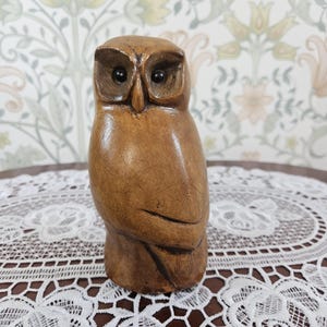 May include: A brown wooden owl figurine with a smooth, polished finish and carved details. The owl features two dark, circular eyes, a pointed beak, and visible wood grain textures throughout its body, standing in an upright, alert posture.