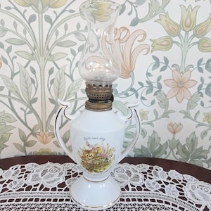 May include: A white porcelain oil lamp with two handles and gold trim. The base features a colourful illustration of a small animal in a garden with the text Each new day. A clear glass chimney sits atop the brass burner assembly.