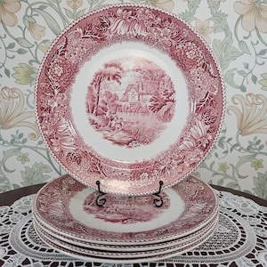 May include: A set of four round ceramic dinner plates featuring a deep red transferware pattern. The centre of each plate displays a detailed landscape scene of a house by a river, while the wide rim is decorated with intricate floral motifs.