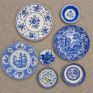May include: A collection of six ceramic plates featuring various blue and white patterns, including floral motifs, landscape scenes, and architectural designs, with one plate displaying a yellow and blue floral pattern.