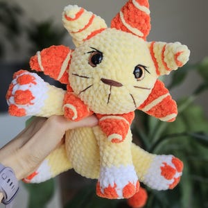 May include: A handmade, plush toy cat in yellow, orange, and white. The cat has large, dark eyes, embroidered whiskers, and orange and white striped accents on its ears and paws. The toy is soft and cuddly.