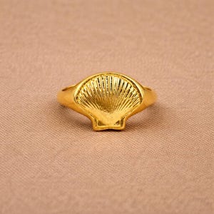 May include: A gold-toned ring featuring a prominent, raised seashell design on the front. The shell motif displays detailed, radiating ridges that extend from the base to the curved outer edge, creating a textured and reflective metallic surface.