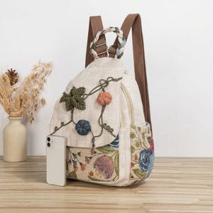 May include: A beige canvas backpack featuring a floral tapestry pattern on the lower section, a braided top handle, and brown adjustable straps. The front flap is decorated with three-dimensional fabric pumpkin and leaf embellishments.