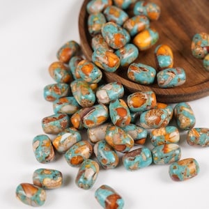 Può includere: Una collezione di perline cilindriche caratterizzate da un motivo marmorizzato nei toni del blu turchese, arancione bruciato e bianco crema. Le perline sono sparse su una superficie, alcune all'interno di un vassoio circolare in legno.