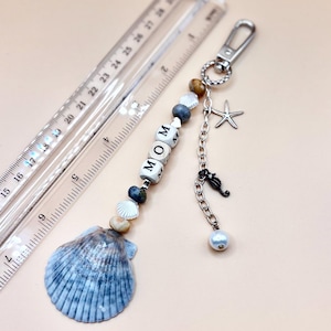 May include: A keychain featuring a large blue and white scallop shell, beads, and white letter cubes spelling MOM. A silver-tone chain includes a starfish charm, a seahorse charm, and a white pearl bead. A clear ruler shows the item is 6 inches long.