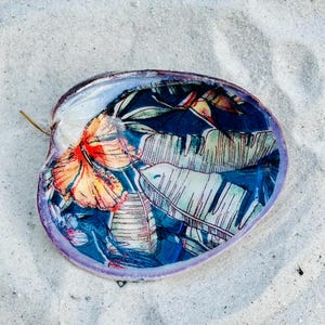 May include: A large seashell with a purple rim, resting on white sand. The interior is decorated with a tropical floral pattern featuring orange hibiscus flowers and green and white leaves against a dark blue background.