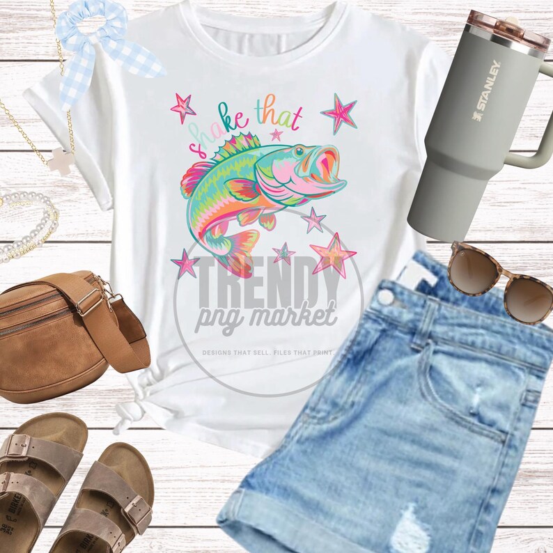 Pu&ograve; includere: T-shirt bianca con grafica di pesce colorata e la scritta "Shake That". L'immagine include anche pantaloncini di jeans, occhiali da sole, un bicchiere Stanley, sandali, un elastico per capelli e una borsa a tracolla marrone.