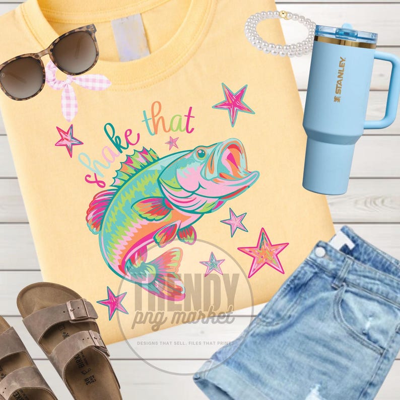 Pu&ograve; includere: Felpa gialla con grafica di pesce colorata e la scritta "Shake that". Occhiali da sole, un bicchiere Stanley azzurro, pantaloncini di jeans, sandali e un braccialetto di perle sono anche nell'immagine.
