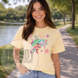 Pu&ograve; includere: T-shirt giallo chiaro con una grafica di pesce colorato e la scritta "be that", pi&ugrave; stelle. Il pesce ha scaglie verdi, rosa e blu. Abbigliamento casual.