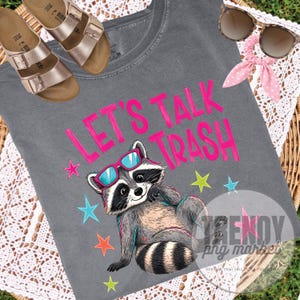 Pu&ograve; includere: T-shirt grigio con la scritta rosa "LET'S TALK TRASH" e un procione dei cartoni animati con occhiali da sole. La maglietta &egrave; decorata con stelle colorate. Sono visibili anche sandali dorati e occhiali da sole con un fiocco rosa.