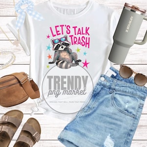Pu&ograve; includere: T-shirt bianca con grafica di procione con occhiali da sole e la scritta "LET'S TALK TRASH". L'immagine include anche pantaloncini di jeans, un bicchiere Stanley grigio, occhiali da sole e altri accessori.