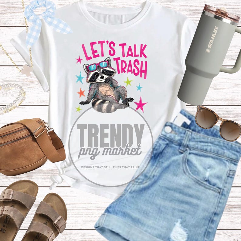 Pu&ograve; includere: T-shirt bianca con grafica di procione con occhiali da sole e la scritta "LET'S TALK TRASH". L'immagine include anche pantaloncini di jeans, un bicchiere Stanley grigio, occhiali da sole e altri accessori.
