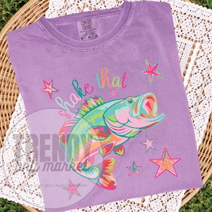 Pu&ograve; includere: Una maglietta lilla Comfort Colors con una grafica colorata di un pesce con la bocca aperta, circondato da stelle. Il testo "shake that" &egrave; scritto sopra il pesce in un carattere giocoso. La maglietta &egrave; esposta su una superficie tessuta.