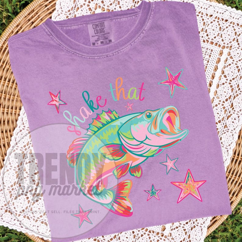 Pu&ograve; includere: Una maglietta lilla Comfort Colors con una grafica colorata di un pesce con la bocca aperta, circondato da stelle. Il testo "shake that" &egrave; scritto sopra il pesce in un carattere giocoso. La maglietta &egrave; esposta su una superficie tessuta.