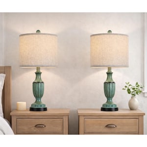 May include: Two matching table lamps with teal bases and beige fabric shades. The lamps sit on wooden nightstands, with a candle and a small vase of greenery nearby. The lamps are turned on, illuminating the room.