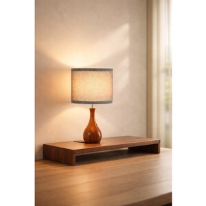May include: A wooden desk lamp with a brown, teardrop-shaped base and a gray lampshade. The lamp sits on a matching wooden monitor stand. The lamp is turned on, illuminating the room. The desk and stand are made of wood.
