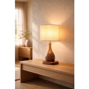 May include: A wooden table lamp with a beige linen shade. The lamp has a teardrop-shaped wooden base and sits on a rectangular wooden base. The lamp is on a wooden table in a room with natural light.