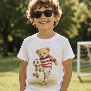 May include: A white short-sleeved t-shirt featuring a graphic print of a teddy bear wearing a red and white striped soccer jersey with the number 10 on the shorts, white socks with blue and red stripes, and yellow cleats while kicking a soccer ball.