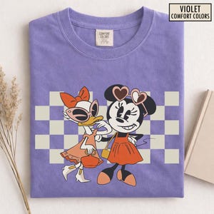 Pu&ograve; includere: T-shirt Comfort Colors viola con un'illustrazione di Minnie Mouse e Paperina. Minnie Mouse indossa occhiali da sole a forma di cuore e un vestito arancione. Paperina indossa occhiali da sole e un vestito arancione. La maglietta ha uno sfondo a scacchi.