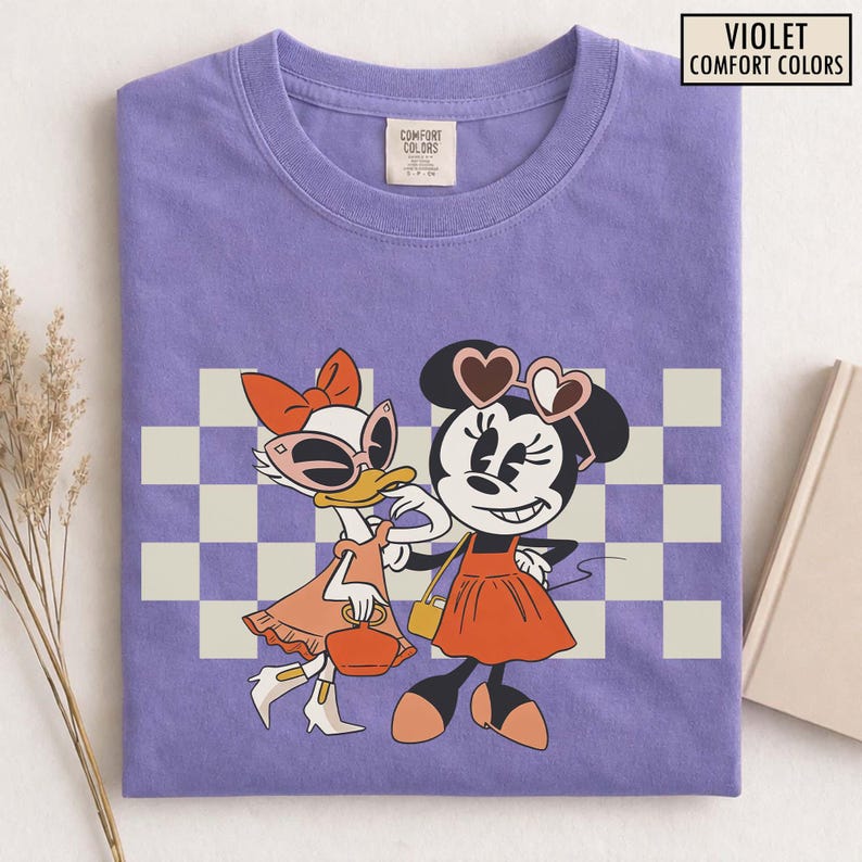 Pu&ograve; includere: T-shirt Comfort Colors viola con un'illustrazione di Minnie Mouse e Paperina. Minnie Mouse indossa occhiali da sole a forma di cuore e un vestito arancione. Paperina indossa occhiali da sole e un vestito arancione. La maglietta ha uno sfondo a scacchi.