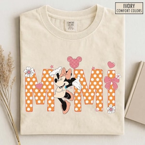 May include: Ivory Comfort Colors t-shirt featuring the word "MIMI" in orange polka dot letters, with a Minnie Mouse graphic. The design includes pink hearts, stars, and flowers. The shirt is folded.
