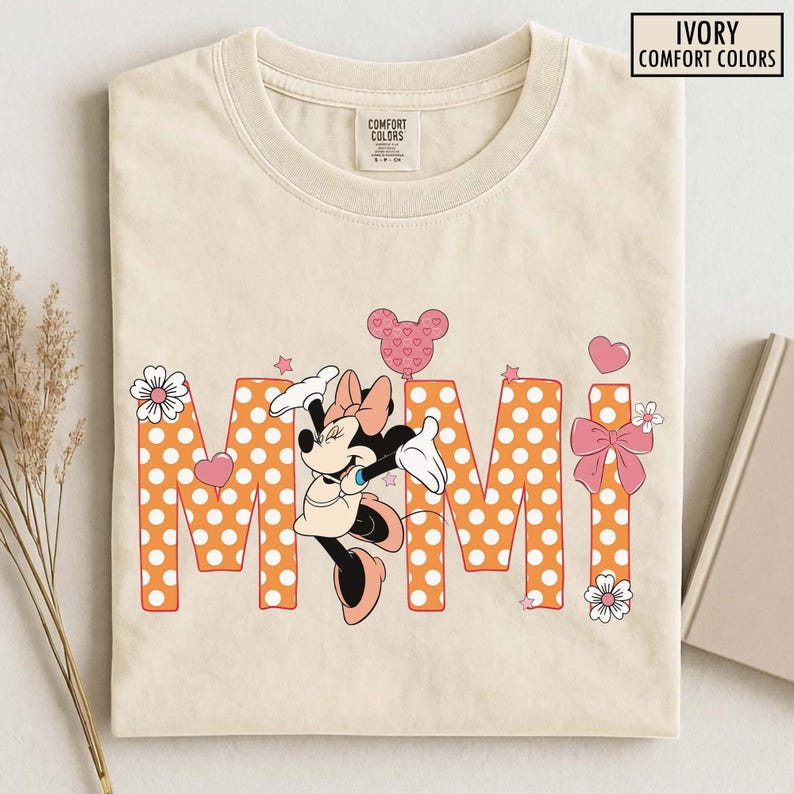 May include: Ivory Comfort Colors t-shirt featuring the word "MIMI" in orange polka dot letters, with a Minnie Mouse graphic. The design includes pink hearts, stars, and flowers. The shirt is folded.