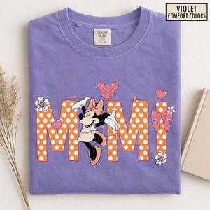 May include: Violet Comfort Colors t-shirt featuring a Minnie Mouse graphic. The word "Mimi" is spelled out in orange polka dot letters. The shirt has a crew neck and a small Comfort Colors tag.
