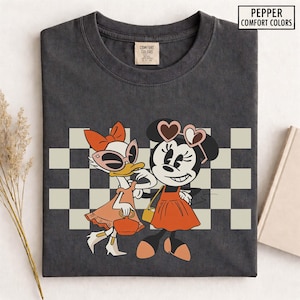 Pu&ograve; includere: T-shirt grigio scuro con un'illustrazione a cartoni animati di Paperina e Minnie. Paperina indossa occhiali da sole e un vestito, mentre Minnie indossa occhiali da sole a forma di cuore e un vestito. Lo sfondo ha un motivo a scacchi. Il testo "PEPPER COMFORT COLORS" &egrave; in alto a destra.