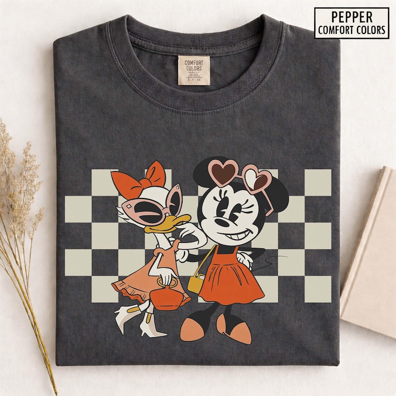 Pu&ograve; includere: T-shirt grigio scuro con un'illustrazione a cartoni animati di Paperina e Minnie. Paperina indossa occhiali da sole e un vestito, mentre Minnie indossa occhiali da sole a forma di cuore e un vestito. Lo sfondo ha un motivo a scacchi. Il testo "PEPPER COMFORT COLORS" &egrave; in alto a destra.